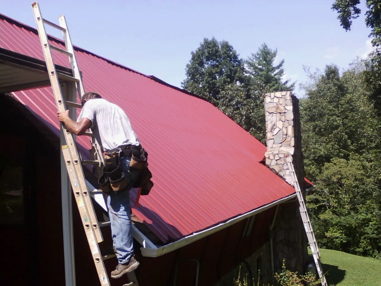 Roofer on a ladder beside a red metal roof and stone chimney — Soffit & Fascia Repair by Roofing Brothers in Studio City